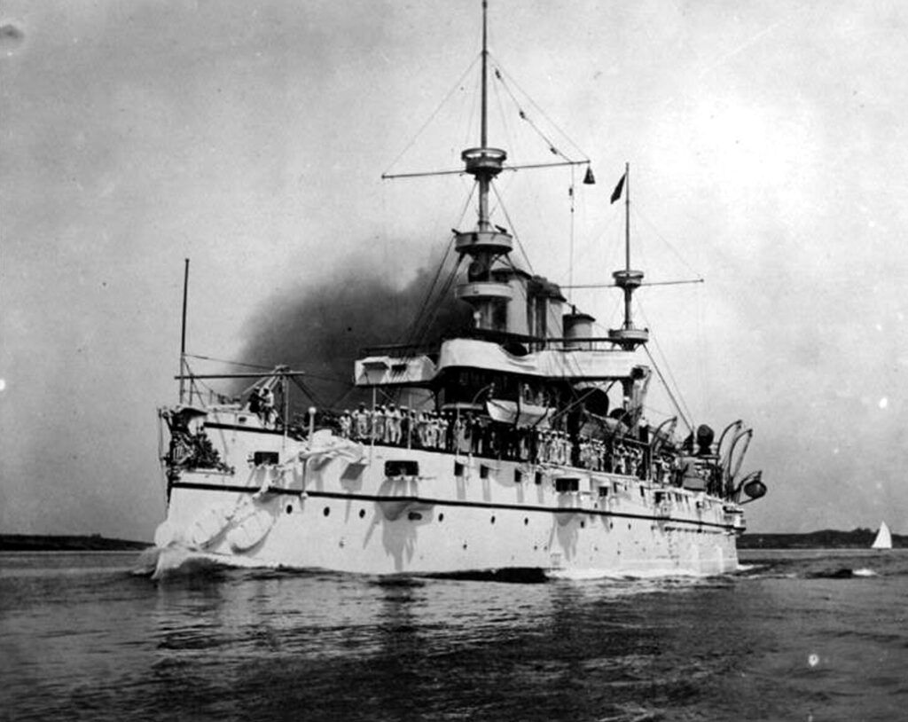 A black and white photograph of the USS New York (ACR-2), an early 20th-century armored cruiser, steaming across the water with a large crew visible on deck and smoke billowing from its smokestack.