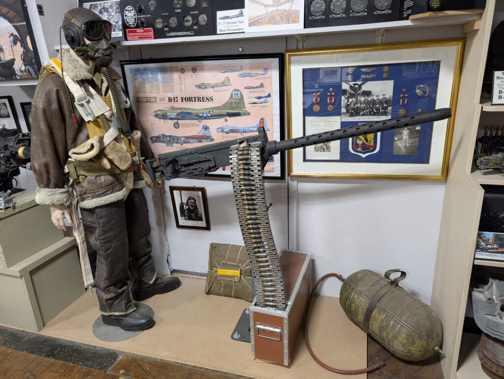 A museum exhibit featuring a mannequin in a WWII bomber crewman's flight gear standing next to a Browning M2 machine gun. In the background are B-17 Fortress memorabilia.
