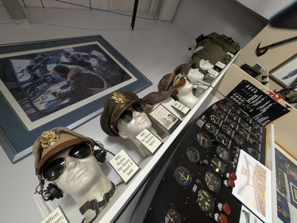 A museum exhibit of World War II U.S. Army Air Forces flight gear, including officer hats, aviator sunglasses, and flight helmets displayed on mannequin heads next to an aircraft instrument panel.