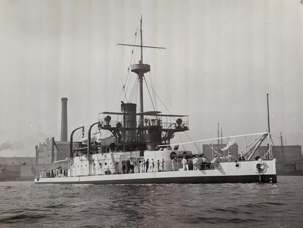 A sepia-toned historical photograph of the U.S. Navy monitor USS Terror (BM-4) at anchor in a harbor, circa 1890s. The ship has a white hull and sailors are visible on deck.