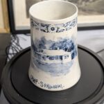 A white ceramic mug with blue hand-painted scenes and the inscription 'CAMP-SORGHUM-' at its base.