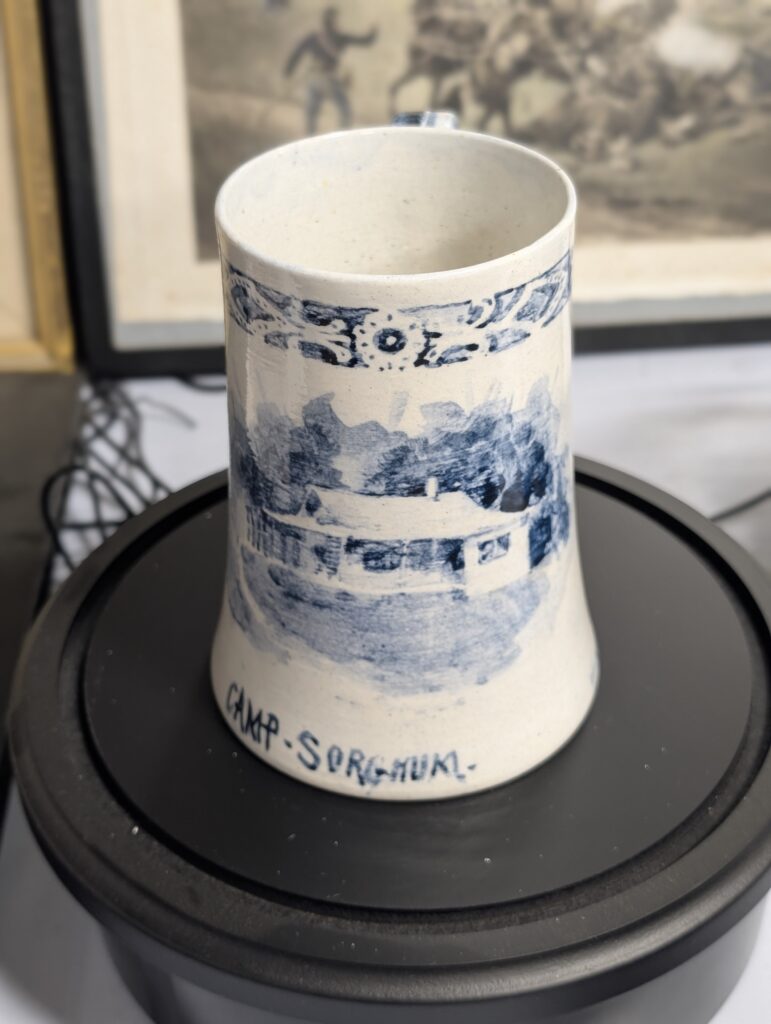 A white ceramic mug with blue hand-painted scenes and the inscription 'CAMP-SORGHUM-' at its base.