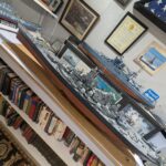 A detailed display of military ship models, framed certificates, and books, featuring models of the USS Arizona battleship and the USS Hornet aircraft carrier, set in what appears to be a personal military history collection.