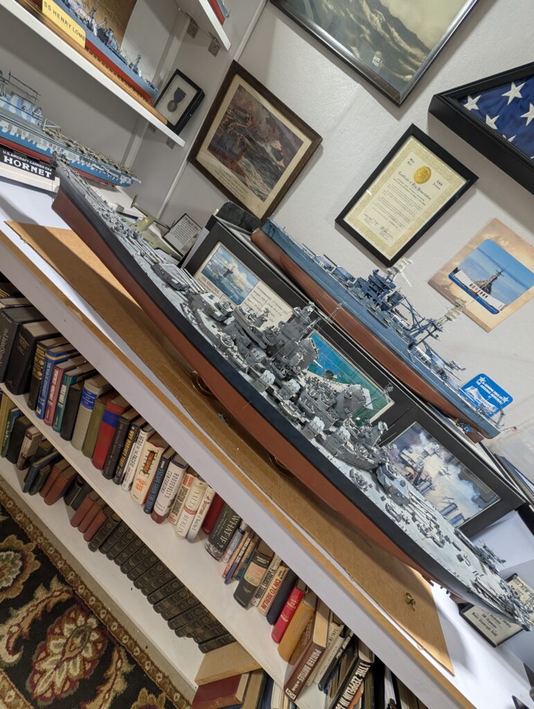 A detailed display of military ship models, framed certificates, and books, featuring models of the USS Arizona battleship and the USS Hornet aircraft carrier, set in what appears to be a personal military history collection.