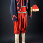 A full military uniform on display, featuring a dark blue jacket with red trim and ornate red designs, red baggy pants, white stockings, and black shoes. A red fez with a yellow tassel is placed next to the mannequin.